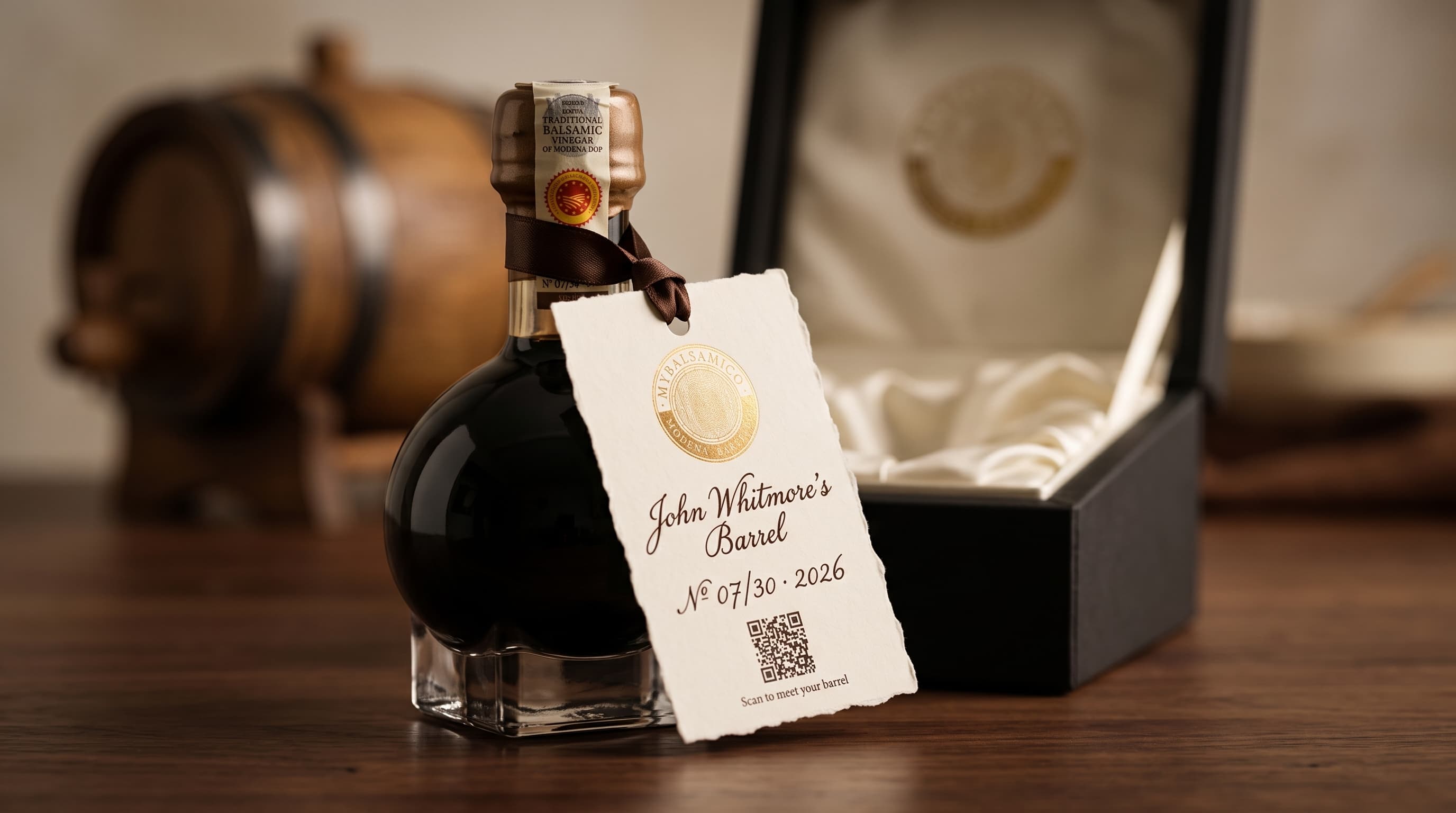 Boccetta di aceto balsamico tradizionale con il cartellino «John Whitmore's Barrel · N° 07/30 · 2026», il QR code e, sullo sfondo, una piccola botte e il cofanetto aperto